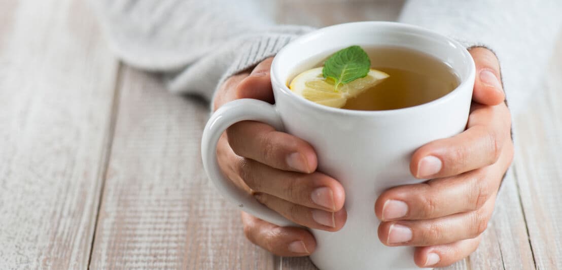 Zwei Hände, die eine weiße Tasse halten, die auf einem Tisch steht. In der Tasse befindet sich Tee, eine Scheibe Zitrone und ein grünes Kräuterblatt.