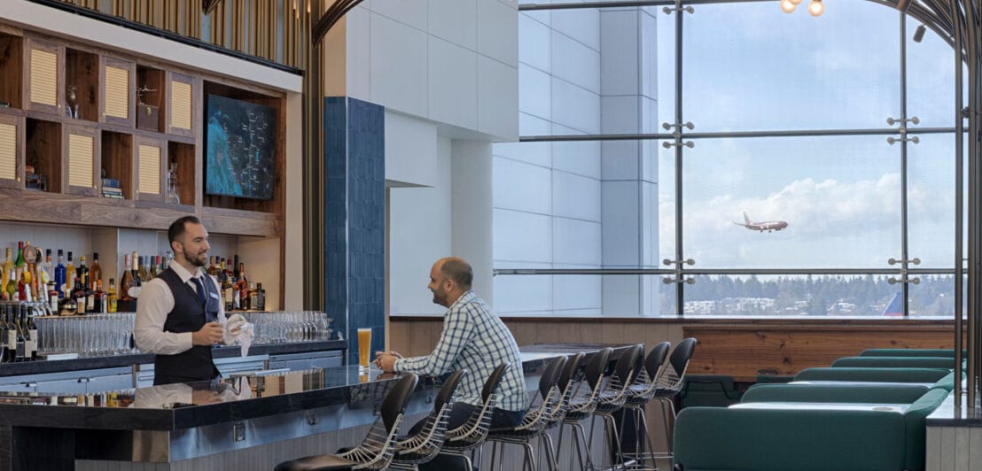Ein Mann sitzt an der Bar einer modernen Flughafen-Lounge mit Blick aufs Rollfeld.