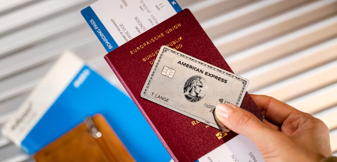 Eine silberne Kreditkarte von American Express auf einem Reisepass mit Bordkarte, gehalten von einer Hand.