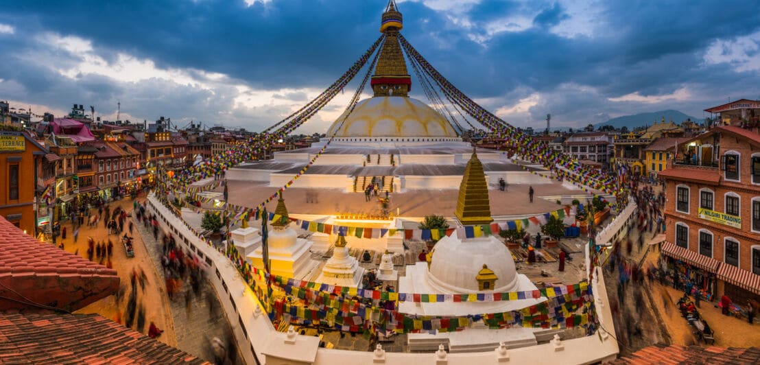 Buddhistische Tempelanlage mit weiß-goldener Kuppel und tibetischen Gebetsflaggen in einem Stadtkern mit Menschen.