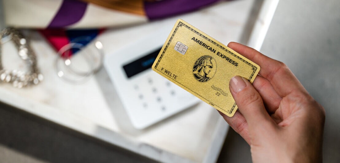 Eine Hand hält eine goldene Kreditkarte von American Express über ein Kartenlesegerät neben Modeaccessoires.