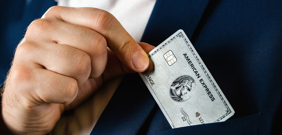 Eine Hand steckt eine silberne Kreditkarte von American Express in die Reverstasche eines blauen Jacketts.