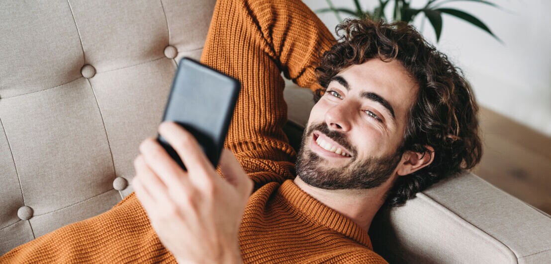 Ein junger Mann liegt entspannt auf dem Sofa und schaut lächelnd auf sein Smartphone.