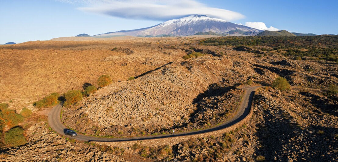 Ein Auto fährt auf einer kurvigen Landstraße in karger Landschaft, im Hintergrund ein schneebedeckter Vulkan.