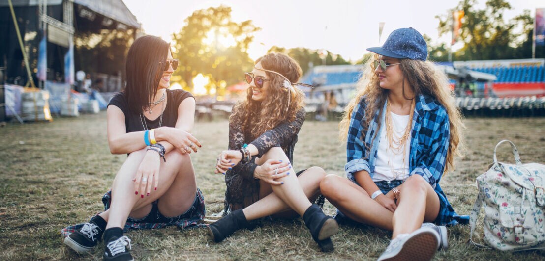 Drei Frauen sitzen auf einem Festivalgelände, hinter ihnen die Bühne.