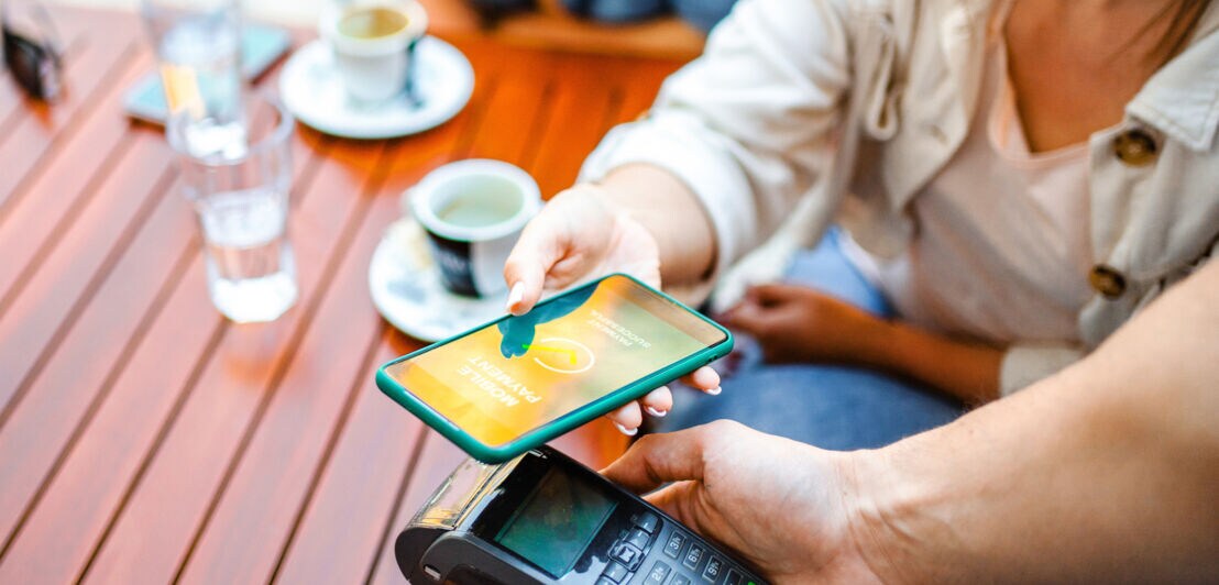 Nahaufnahme einer kontaktlosen Zahlung mit Kreditkarte via Smartphone an einem Kartenlesegerät an einem Cafétisch im Freien.