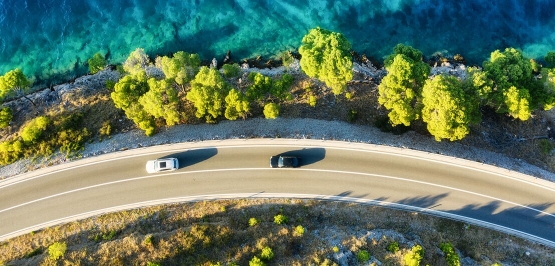 Zwei Autos fahren auf einer Küstenstraße entlang grüner Bäume am türkisblauen Meer bei tiefem Sonnenschein aus der Vogelperspektive.