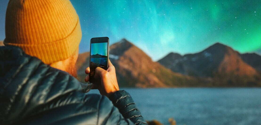 Ein Mann mit gelber Mütze hält ein Smartphone in der Hand und fotografiert damit Polarlichter.