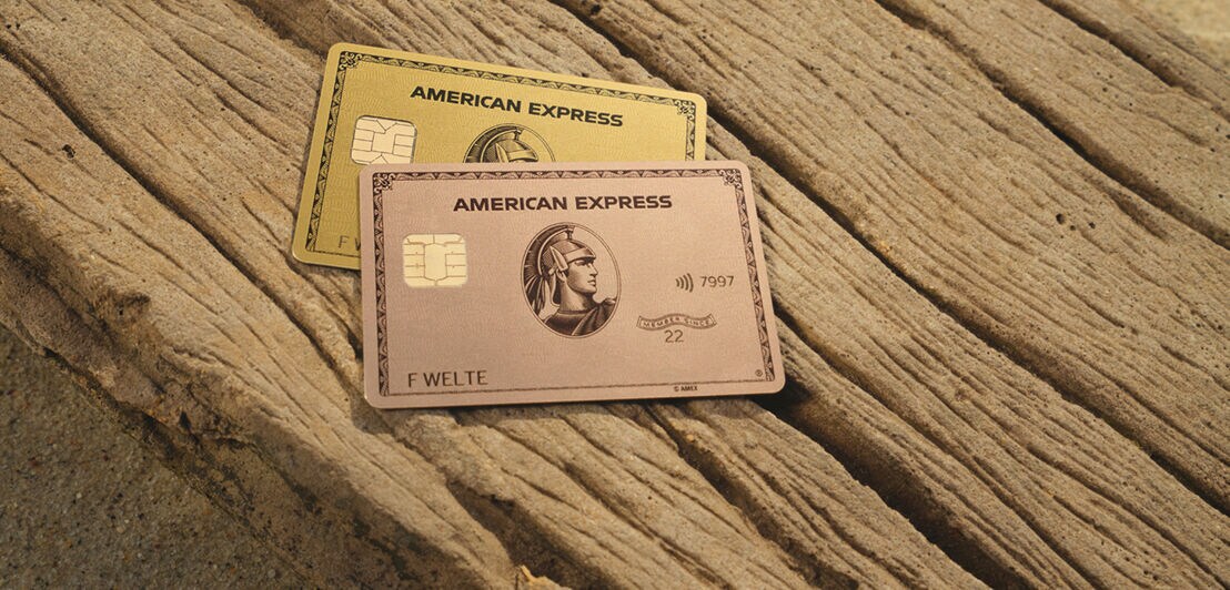 Eine goldene und eine rosé-goldene Kreditkarte von American Express liegen bei Sonnenschein auf einem Stück Treibholz.