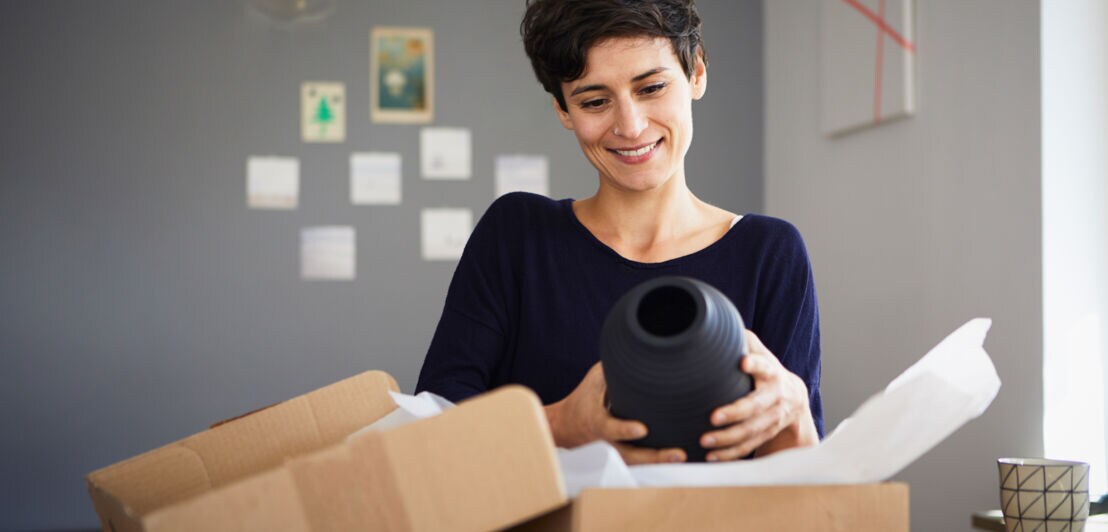 Eine lächelnde Frau hält eine schwarze Vase über ein Paket mit Packpapier auf einem Tisch.