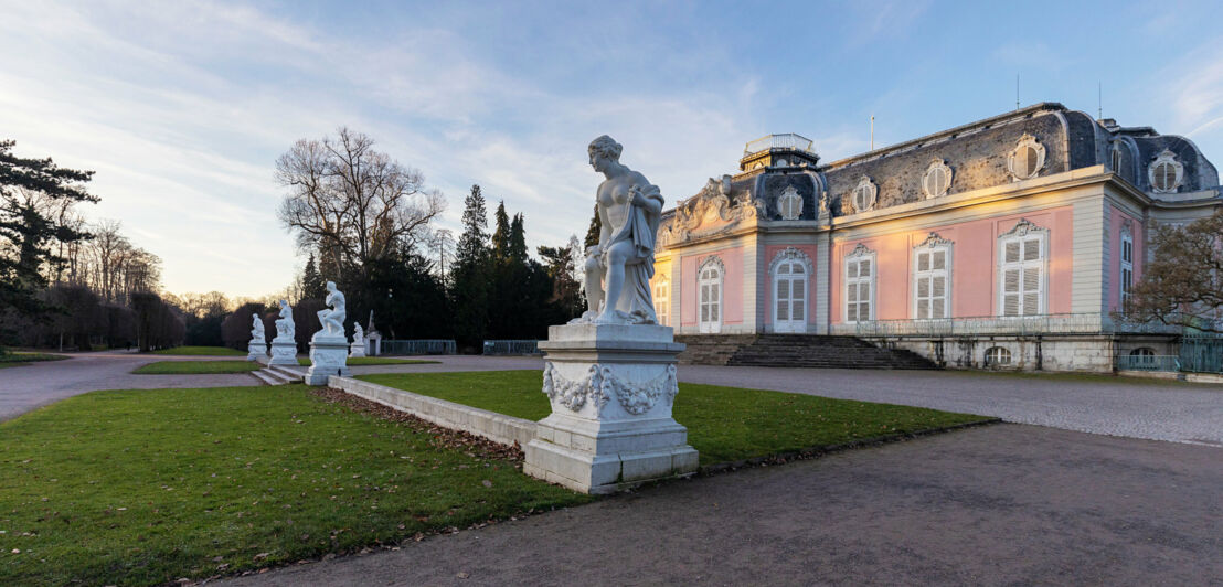 Rosafarbenes Schloss mit Steinskulpturen in einer Parkanlage im Vordergrund.