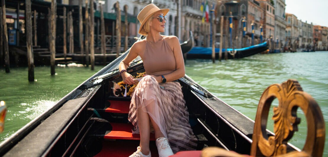 Eine Frau mit Sonnenbrille und Sonnenhut sitzt in einer Gondel auf einem Kanal in Venedig.