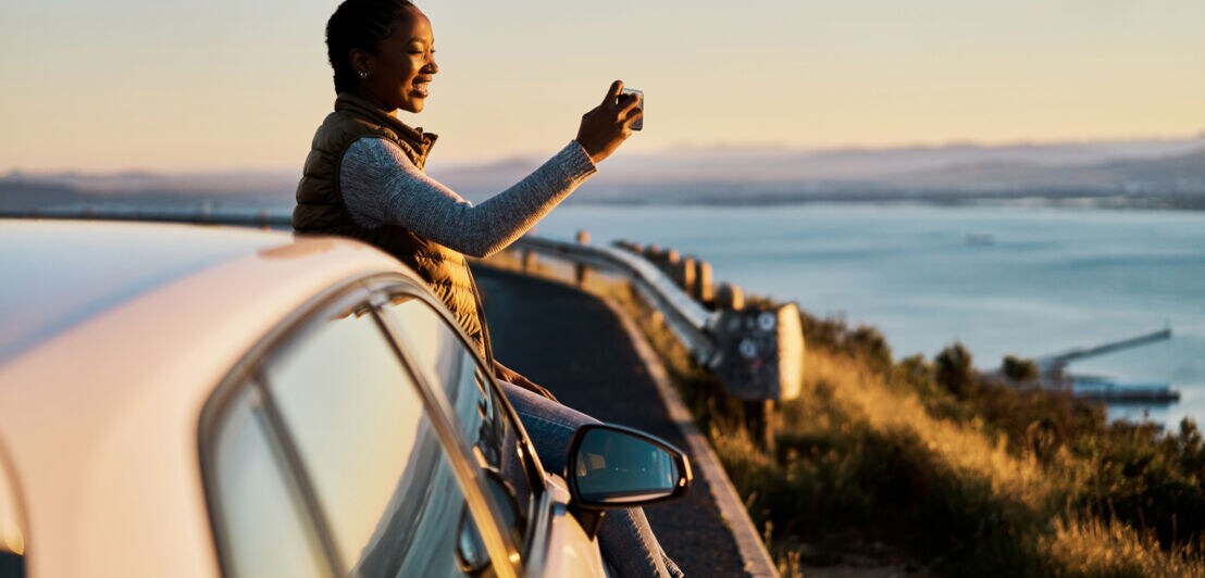 Eine Frau sitzt auf der Motorhaube eines parkenden Autos auf einer Küstenstraße und macht ein Foto mit ihrem Smartphone.