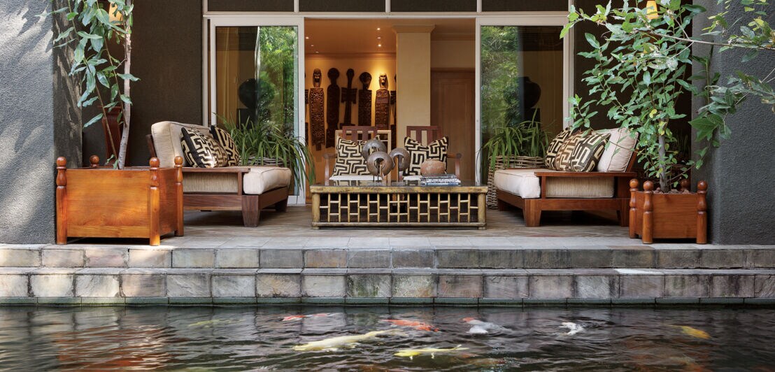Terrasse mit Holzmöbeln und Kunstwerken im afrikanischen Stil an einem Wasserbecken mit Koikarpfen.