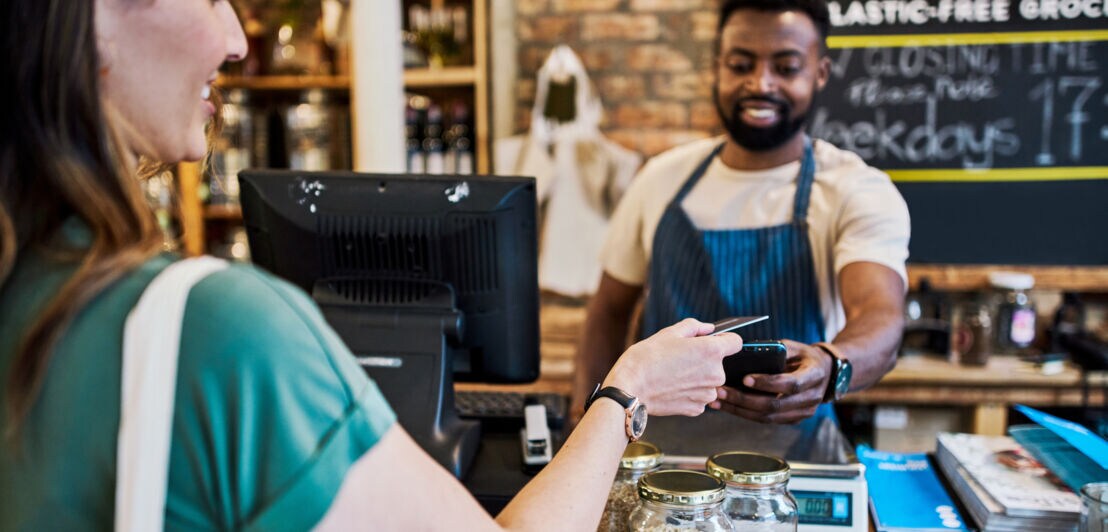 Eine Frau bezahlt kontaktlos mit ihrer Kreditkarte an der Theke in einem Geschäft, der Verkäufer hält ihr ein Kartenlesegerät entgegen.