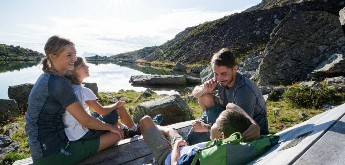 Eine Familie mit zwei Kindern in Wanderkleidung sitzt auf einer Holzbank an einem See in den Bergen.