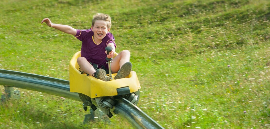 Ein fröhlicher Junge fährt in einem Bobschlitten auf einer Schiene über eine grüne Wiese.