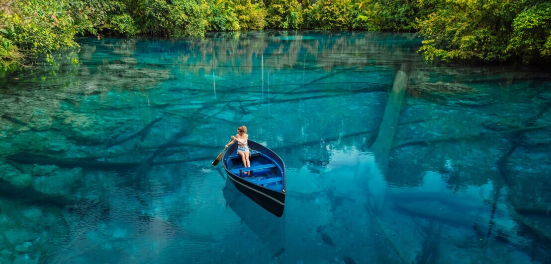 Eine Frau sitzt in einem Ruderboot auf einem blauen, glasklaren See, umgeben von grüner Vegetation.