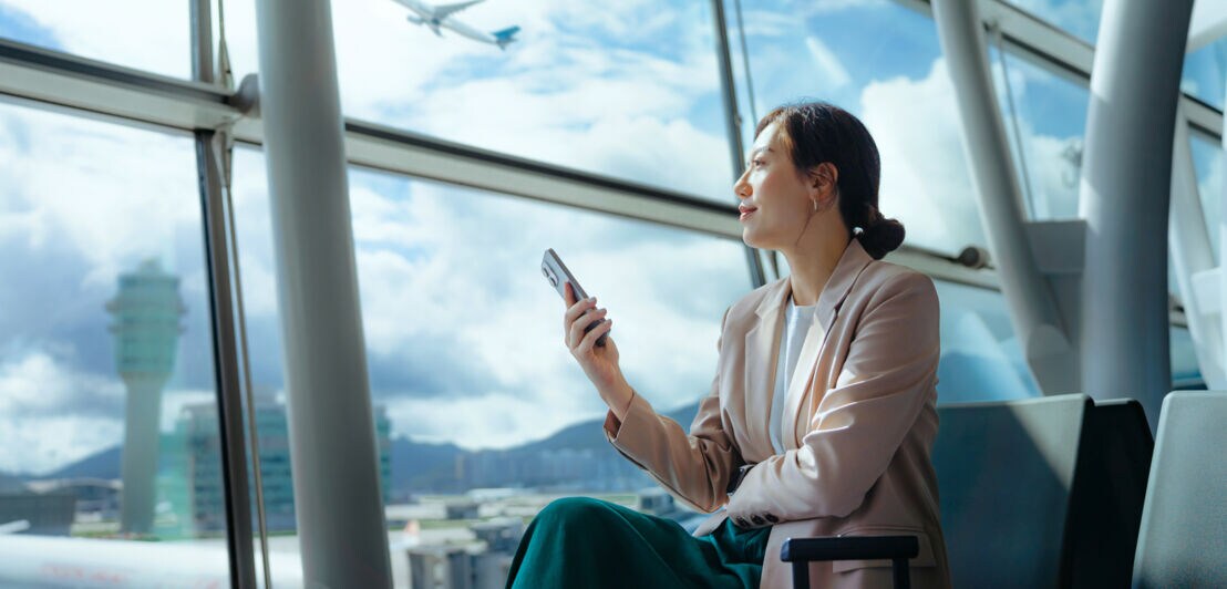 Eine moderne Geschäftsfrau sitzt mit einem Smartphone in ihrer Hand an einem Gate am Flughafen und blickt aufs Rollfeld neben ihr steht ein Trolley.