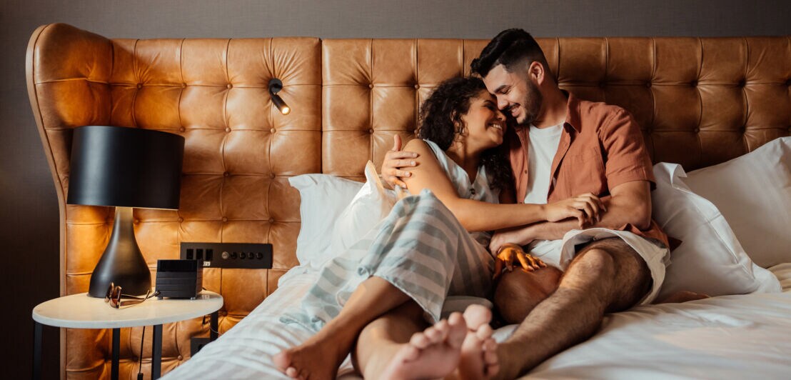 Ein glückliches Paar liegt kuschelnd in einem Hotelbett bei Tageslicht.
