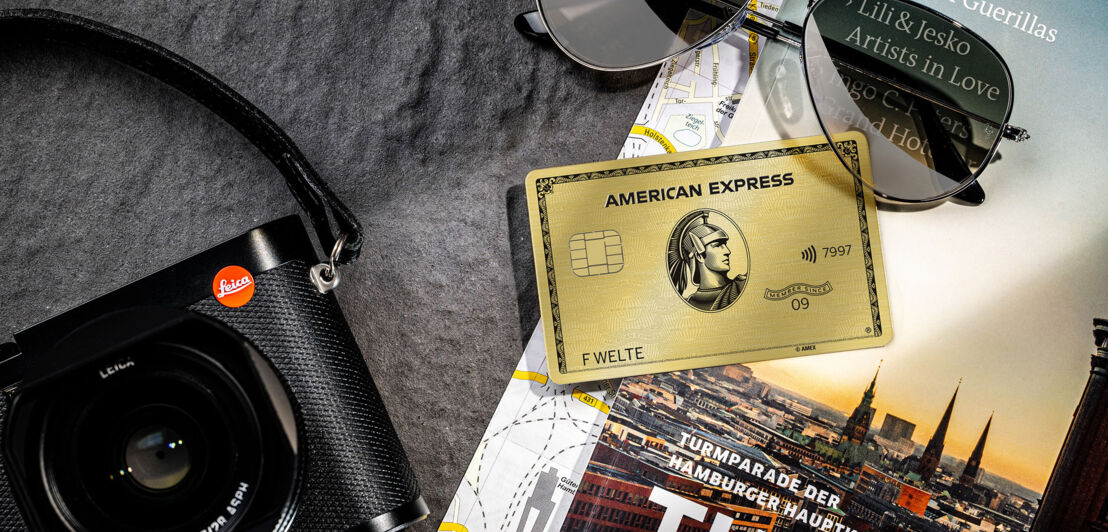 Eine goldene Kreditkarte liegt neben einer Kamera auf einem Reiseführer.