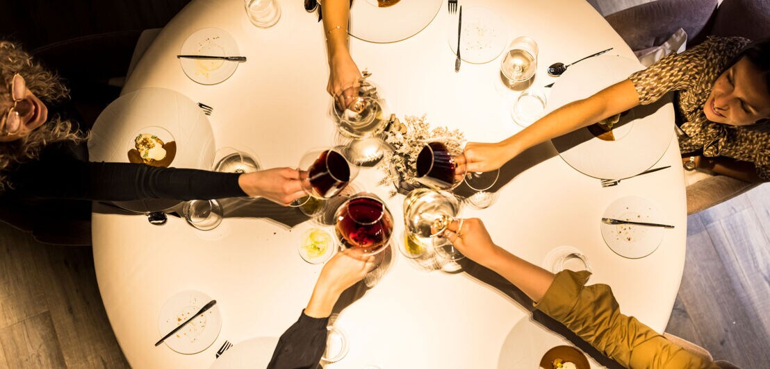 Aufsicht von fünf Armen mit Weingläsern beim Anstoßen über einem runden Esstisch bei dämmrigen Licht.