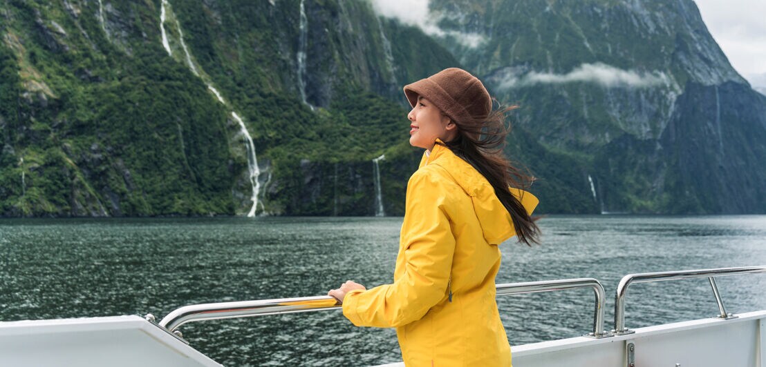 Eine Frau im gelben Regenmantel steht an der Reling eines Schiffes in einer Fjordlandschaft.