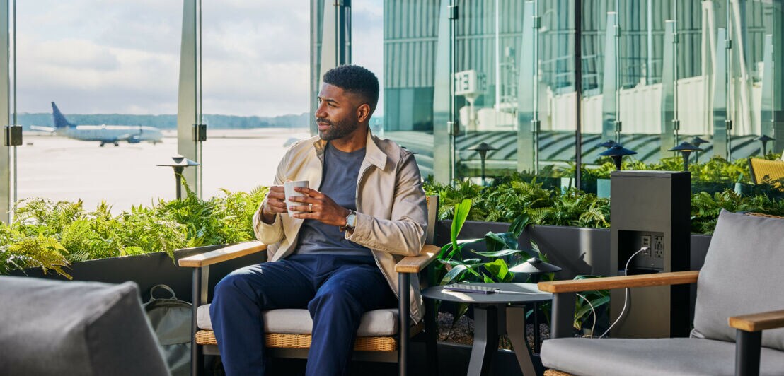 Ein Mann sitzt mit einer Kaffeetasse auf einer Terrasse mit Blick auf ein Flughafenrollfeld.