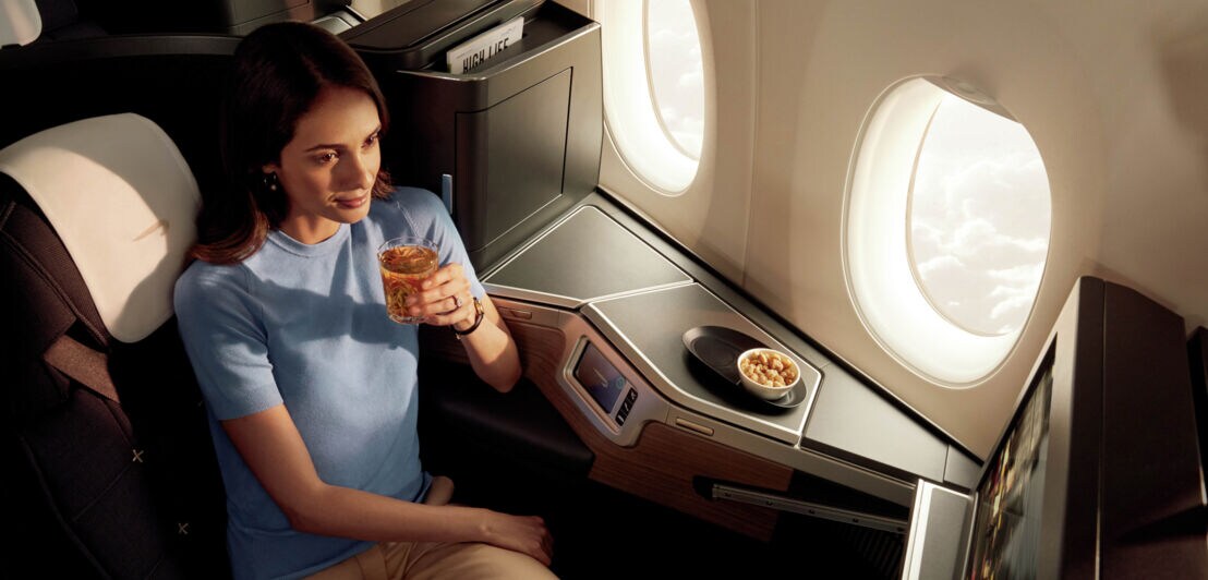 Eine Frau sitzt entspannt mit einem Getränk in der Business Class eines Flugzeugs.