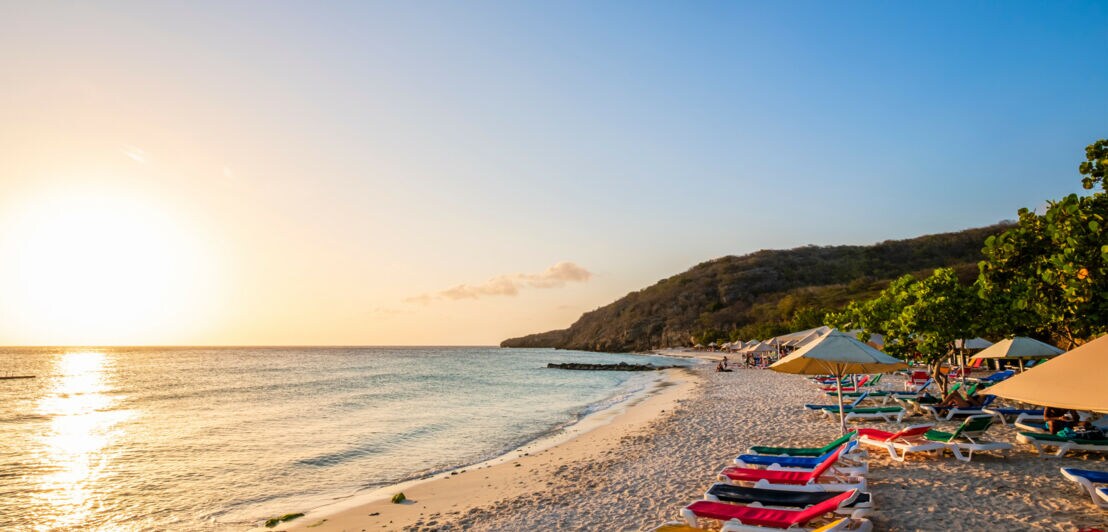 Strand bei Sonnenuntergang mit bunten Liegen und Sonnenschirmen.