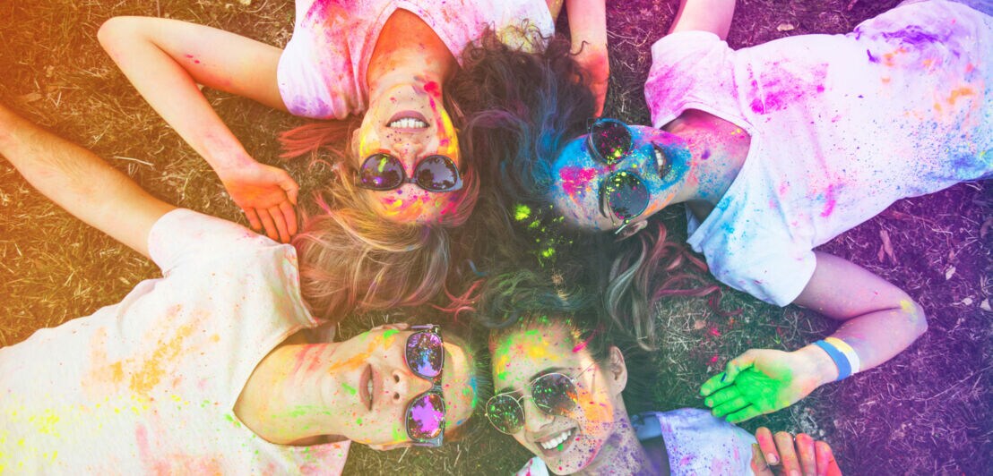 Vier Festivalbesucherinnen mit Sonnenbrillen und farbig besprenkelten Shirts, Gesichtern und Händen liegen lachend auf dem Rasen.