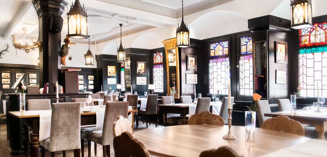 Urig-gemütlicher Innenraum des Restaurants Weinstube mit Buntglasfenstern, edlen Wandvertäfelungen, Rundbögen, verzierter Decke sowie historischen Lampen und Bildern an den Wänden.