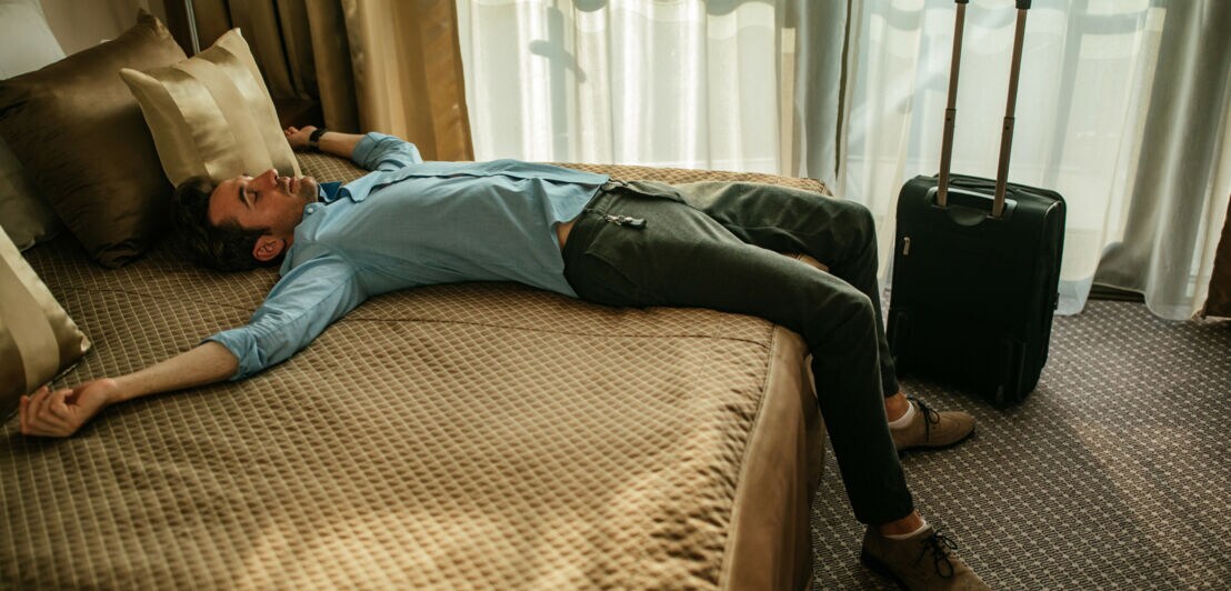 Ein Mann liegt auf dem Rücken mit ausgebreiteten Armen auf einem Hotelbett bei Tageslicht, neben ihm ein Trolley.
