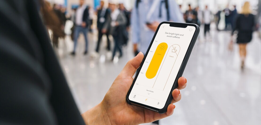 Nahaufnahme eines Smartphones mit Timeshifter App in der Hand einer Person in einem Flughafenterminal.