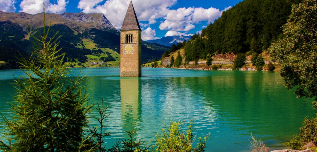 Blick auf den Reschensee, ein malerischer Stausee mit aus dem Wasser ragenden Kirchturm, umgeben von grüner Landschaft.