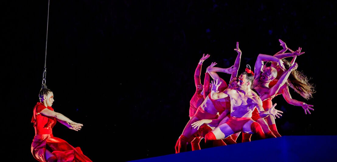 Eine Artistin im roten Kleid an einem Seil vor einer tanzenden Gruppe. 