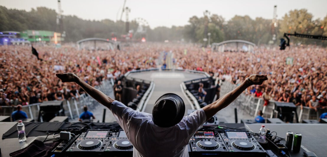 Person von hinten an einem DJ-Pult auf einer Bühne vor einer Zuschauermenge. 