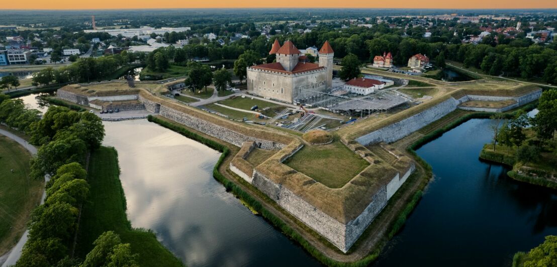 Eine helle Burg mit roten Dächern, umgeben von einem mit Wasser gefüllten Burggraben.