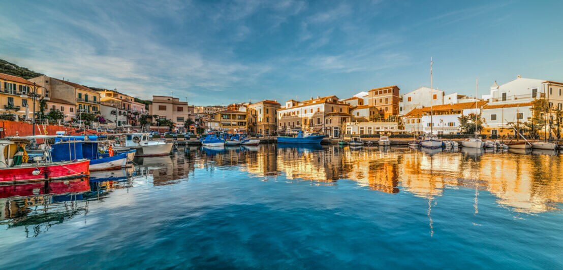 Hafen mit bunten Booten und Gebäuden am Ufer unter bewölktem Himmel, Spiegelung im Wasser.