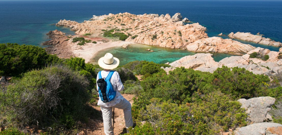 Wandernde Person mit weißem Hut und blauem Rucksack blickt auf felsige Küste und türkisfarbenes Meer.