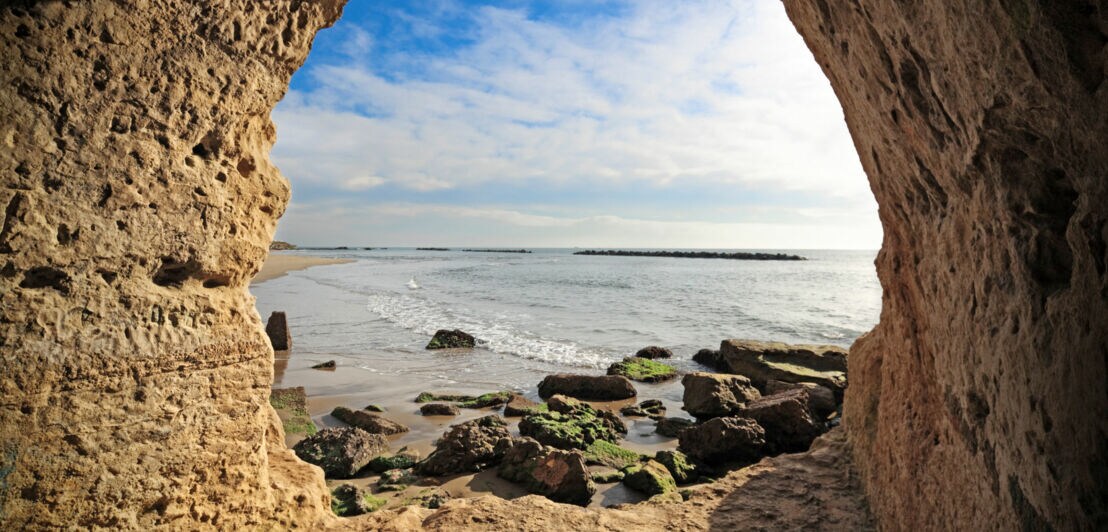 Blick auf einen Sandstrand mit kleinen Felsen im Meer durch eine runde Öffnung in einem großen Felsen am Strand.