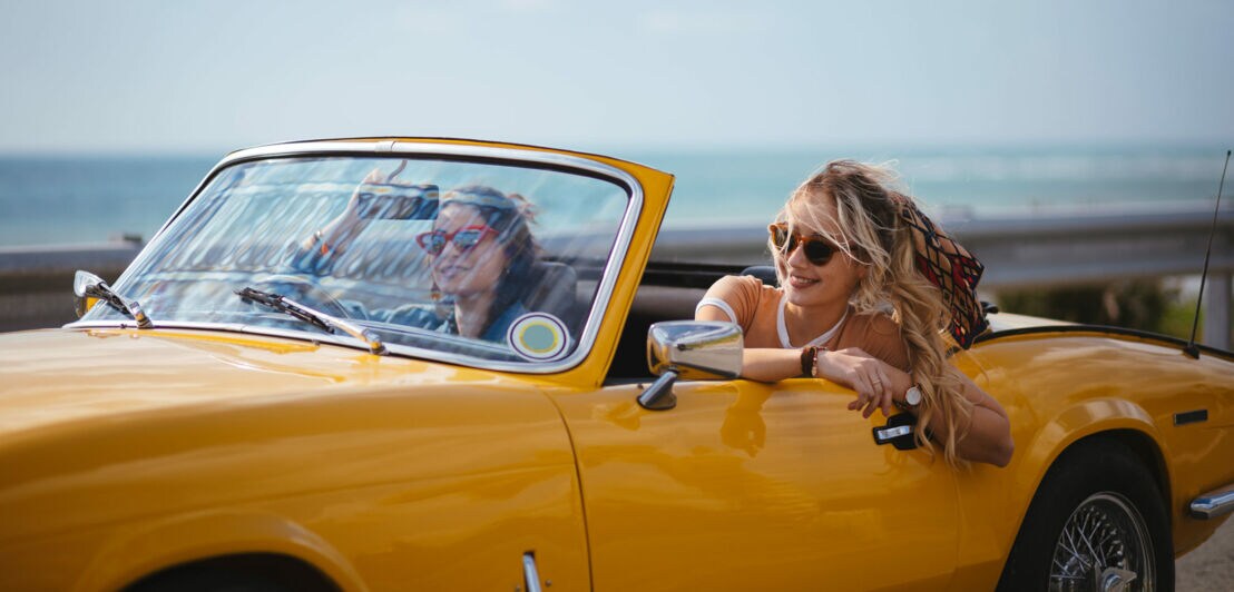 Zwei junge Frauen mit Sonnenbrillen in einem gelben Cabriolet Oldtimer auf einer Küstenstraße.