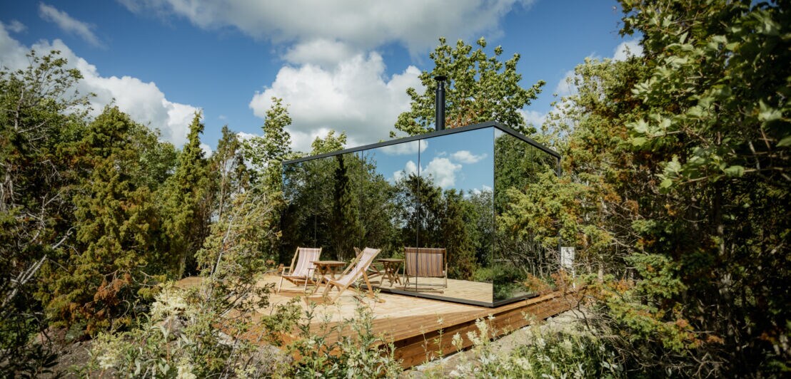 Eine verspiegelte Hütte mit Holzterrasse auf der zwei Liegestühle stehen, umgeben von Laubbäumen und Sträuchern. 