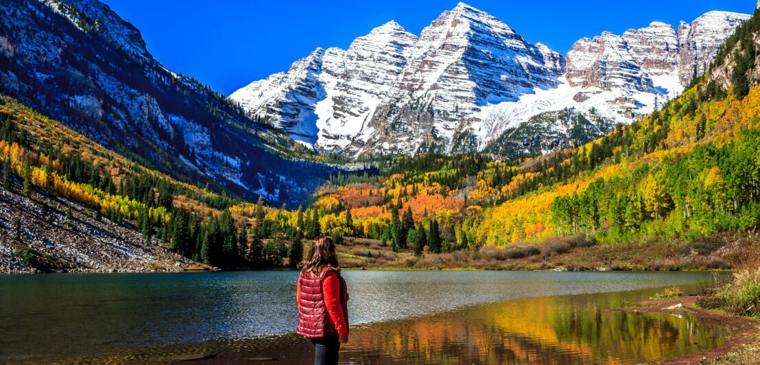 Person in roter Weste steht am Ufer eines Sees mit Blick auf schneebedeckte Berge und herbstlich gefärbten Wald.