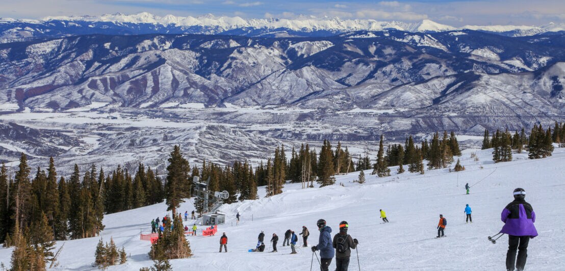 Skifahrende Personen auf schneebedecktem Hang mit Tannenbäumen und Bergen im Hintergrund unter bewölktem Himmel.