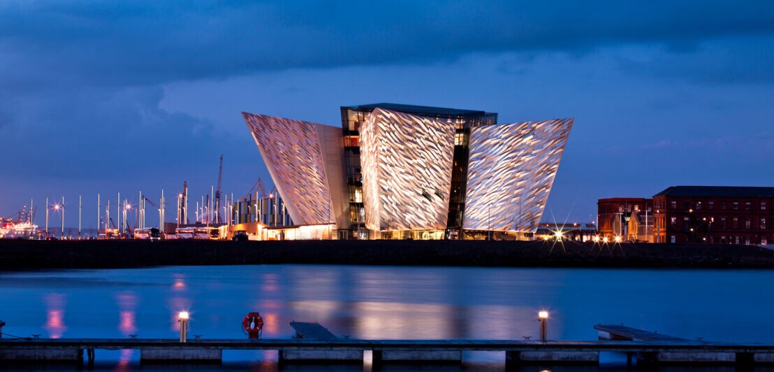 Beleuchtetes modernes Gebäude mit markanter, gefalteter Fassade am Wasser bei Abenddämmerung