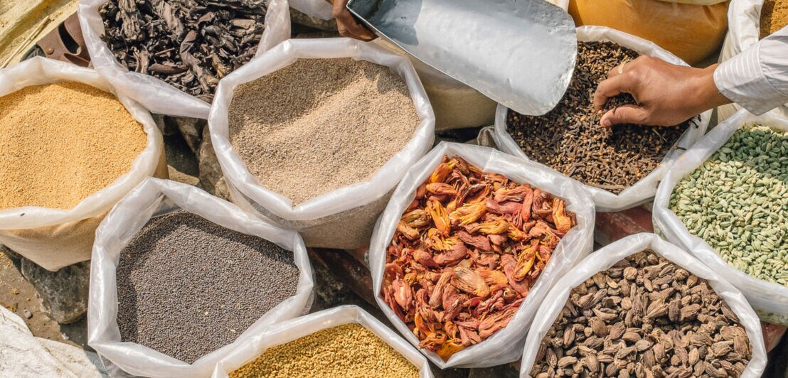 Mehrere Säcke mit verschiedenen getrockneten Gewürzen und Samen auf einem Markt, zwei Hände greifen hinein