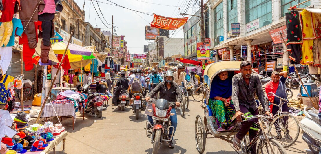 Belebte Straßenmarkt-Szene mit Menschen, Motorrädern, Fahrradrikschas und bunten Ständen in Delhi