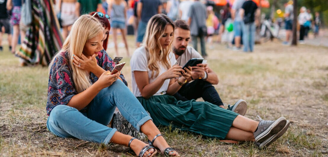 Freunde sitzen im Gras bei einem Festival und schauen auf ihre Smartphones.