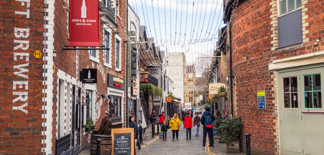 Belebte Kopfsteinpflasterstraße mit Geschäften und Restaurants, darunter eine Craft Brewery, Menschen gehen entlang der Straße unter Lichterketten
